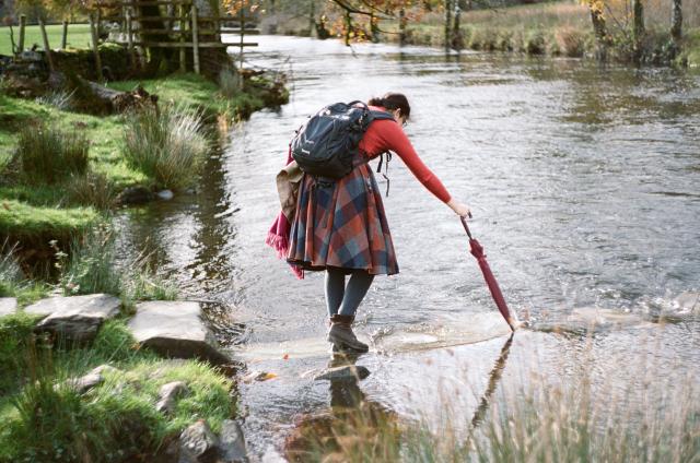 A woman (my wife) dressed in a red sweater and plaid skirt, wearing a black backpack, is crossing a river on stepping stones almost entirely submerged by water. She is using an umbrella to touch the next stone as she leans forward.