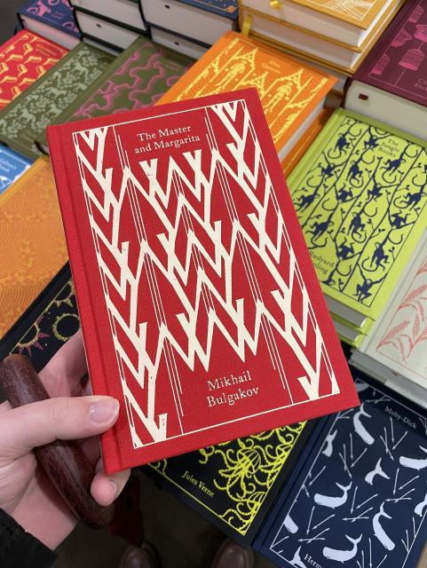 The Master and Margarita by Mikhail Bulgakov as a hardcover book. The cover is red with white lines and triangles. In the background are other books by Jules Verne and Charles Dickens. 