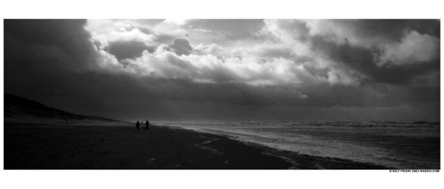 Black and white panorama photograph of a Dutch beach looking southwards at large rain clouds approaching, dunes on the left, North Sea on the right.