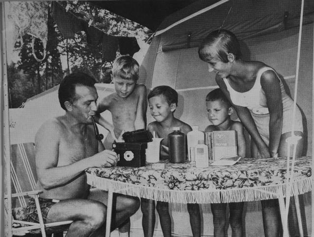 A shirtless man sitting outside a tent and smoking a pipe develops 35mm photographic film using an Agfa Rondinax daylight loading tank. His wife and sons look on with a mixture of admiration and excitement.
