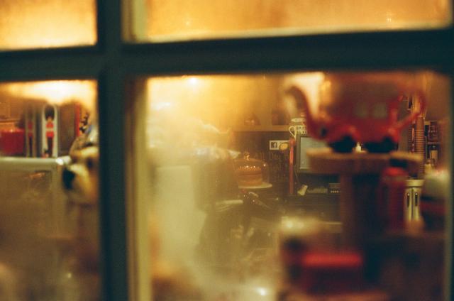 A look through a frosted window onto a space illuminated by amber light and filled by lots of different things, such as a cake under a glass bell, a tea pot shaped like a red double decker bus, a porcelain dog, and many more that cannot be easily identified.
