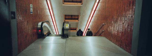 A panoramic photo of steps down to a train platform with an escalator on the left and two men walking down the stairs on the right. The lights have halation around them and the photo has a warm tone