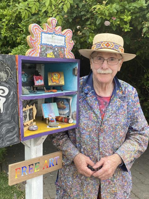 Man in painted coat and panama hat standing next to a little free art gallery.