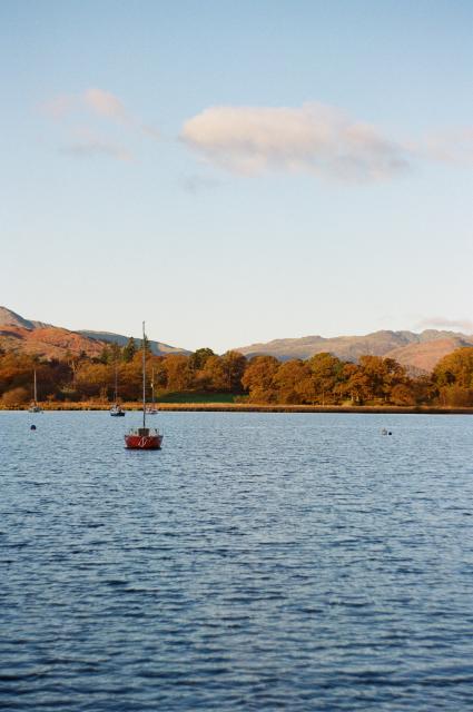 As the sun rises, golden light hits a far away coast filled with autumn colored trees, and behind them, mountains. The sky is pastel blue and mostly cloudless. In the front, a lake that morning light has not yet hit, and close to the middle, a little red boat still coated in darkness.