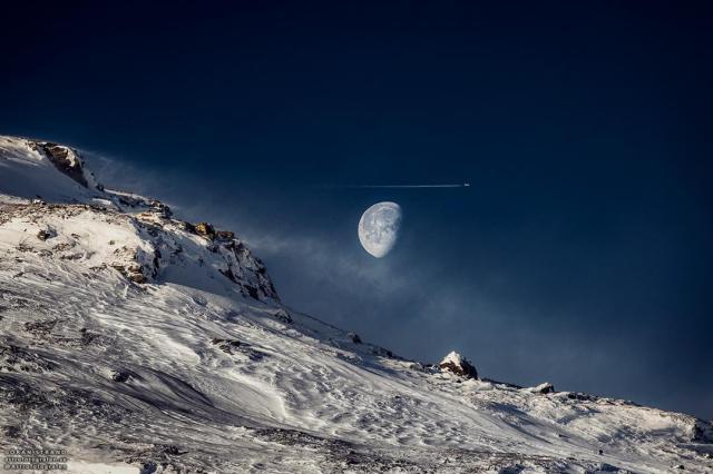 A mostly full moon is seen over a snowy sloping hill. An airplane and contrail are seen just about the Moon.