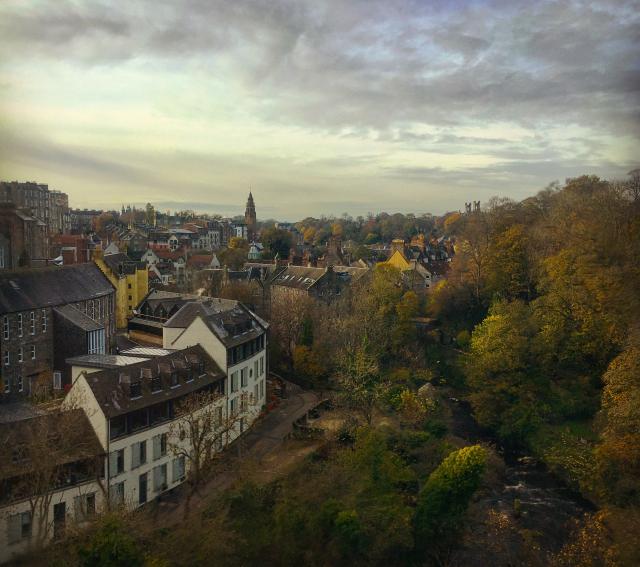Picturesque part of Edinburgh during autumn. Colorful old houses and buildings are stacked around a river bordered by trees and a curved road. The low autumn light and tree colors make the scene atmospheric.