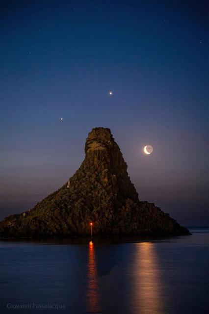 A seascape surrounds a large tree-covered hill. Surrounding the hill in the night sky are three bright dots: the planets Jupiter, Venus, and a crescent Moon.