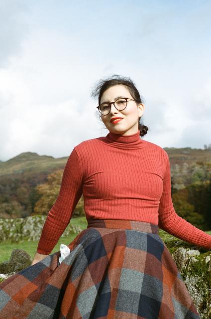 A beautiful woman with black hair and red lips, wearing round glasses, a red turtleneck sweater, and a wool skirt with pockets stands in front of a Northern landscape with mossy rocks, grassy hills, and autumnal trees.