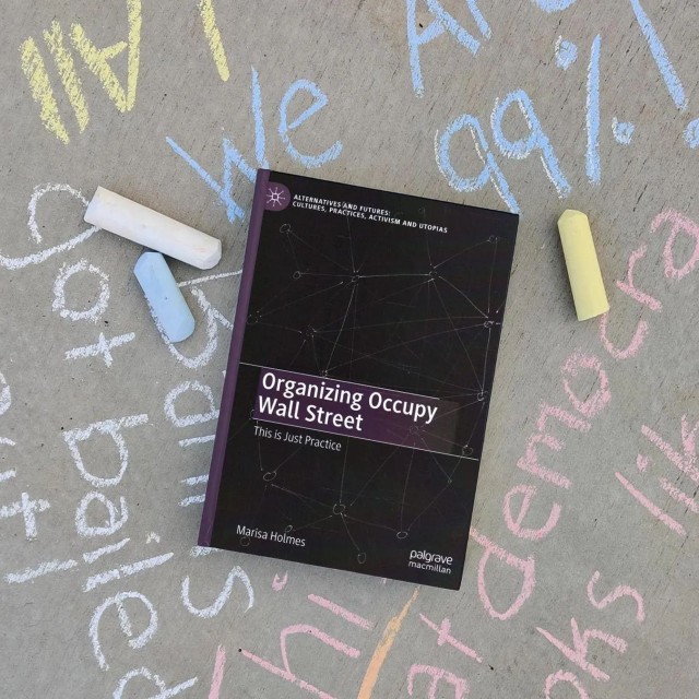 A photo of "Organizing Occupy Wall Street: This is Just Practice" a hardcover book by Marisa Holmes. The book cover is black with a purple spine and features a simple sketch of a network made up of many nodes connected by hastily drawn lines. The book is lying on a sidewalk where slogans have been chalked in several colors, including "We are the 99%!" and "This is what democracy looks like!"