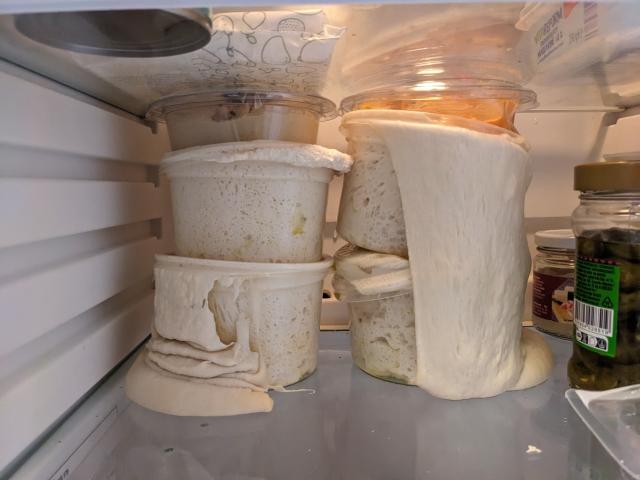 Plastic containers with pizza dough on the fridge are completely spilled over, and have smashed the hummus containers which were placed there to contain the dough.