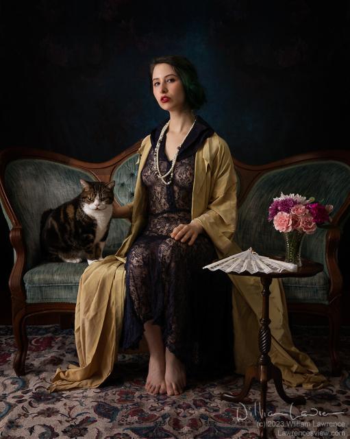 Photography of a woman seated on a settee, wearing a somewhat revealing lace gown, robe, and a pearl necklace.  She is accompanied by a black, brown, and white tabby cat, also seated on the settee.  Both are engaging the camera.  A small table with a lace fan and vase of flowers are in the foreground.  The scene is lit by a soft light from camera left.