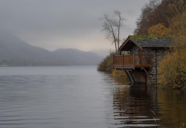 A stone built boathouse with a wooden balcony nestles into the shoreline surrounded by autumnal trees. The lake's water is relatively calm and the view down the lake can be seen. In the background mountains can be made out through the distant mist althought their tops are shrouded by low cloud.