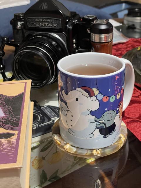 A mug filled with tea on a coaster next to a book. Behind it, a Pentax 6x7 camera. The mug features an elephant shaped snowman wearing a Santa hat being built by little colorful elephants on a snowy landscape. The sky is blurple and decorated with fairy lights. 