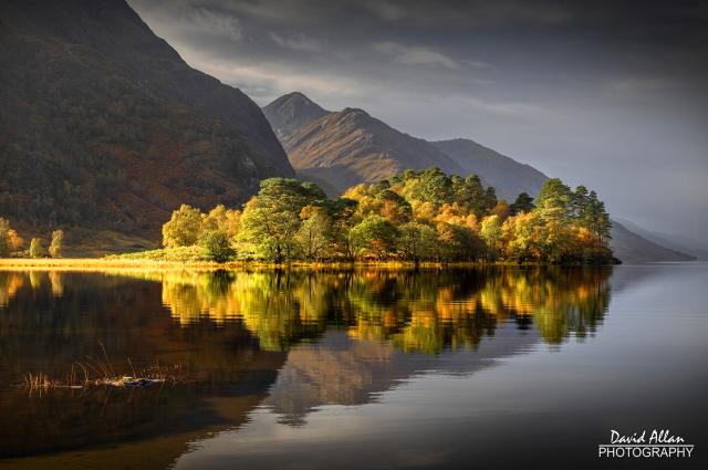 A tree-covered promontory on Loch Shiel at Glenfinnan in the Scottish Western Highlands. The colours of the autumnal season being highlighted by a burst of sunshine.