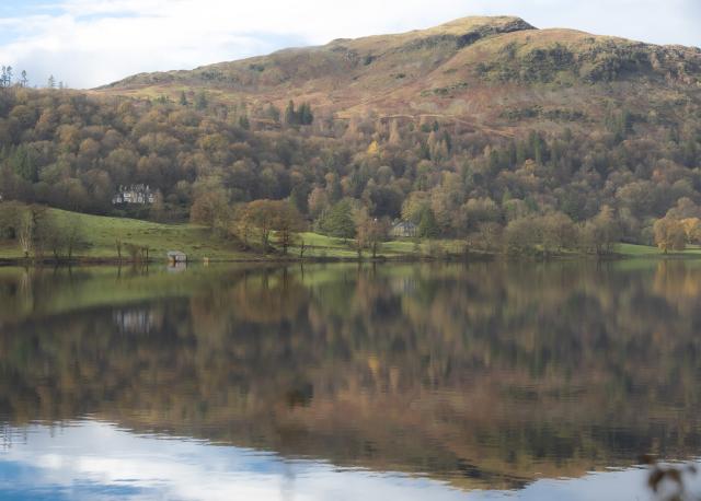 The picture shows a fell (mountain) behind a calm lake. The mountain and the autumn trees are reflected in the water. On the opposite bank is a small boathouse.