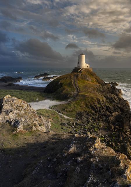 A white painted lighthouse stands on top of a rocky hill. Behind it the sea crashes against the rocks creating lots of white water. The clouds are broken and there is some blue sky visible. The sun is setting to the right of the frame creating shadows and giving the elements in the sun a warm yellow hue