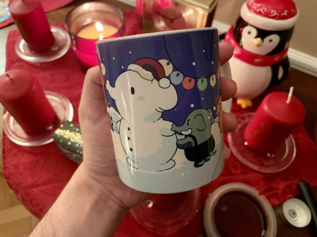 The Mastodon winter mug held in a hand in front of winter decorations, like candles and a penguin shaped cookie jar. The mug itself features little elephants playing in the snow and building an elephant shaped snowman. 