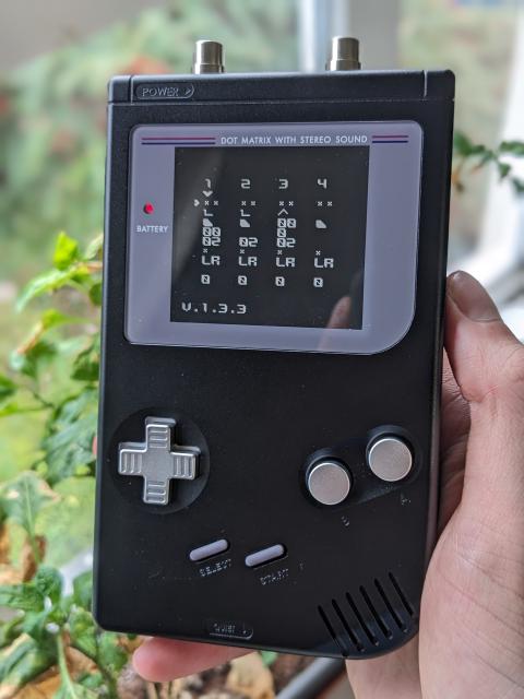 A black anodized aluminum Game Boy with aluminum buttons. A pair of RCA jacks are installed in the top of the Game Boy and it has an IPS display. 