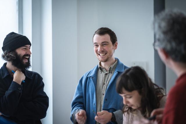 Two people laughing in a daylit room. Two other people out of focus are on the right of the shot.