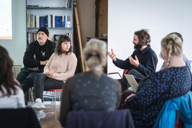 Three members of varia speak animatedly in front of a group of workshop attendees with a small library in the background.