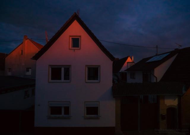 Photograph of houses in a village, against a heavily cloudy, darkening sky, blue with the last light of evening. Peaks of three of the houses are tinged with a pink glow from the setting sun.