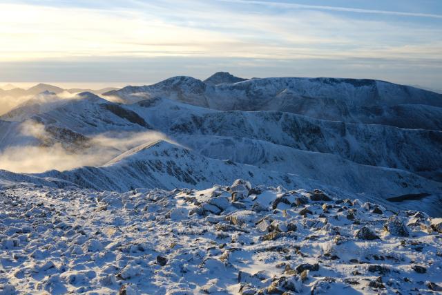 view across a range of snow covered hills and ridges, slight sunlit cloud moving in from the left. the most prominent peak in the distance is ben nevis