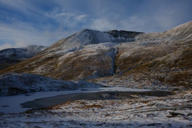 view across a small partially frozen lochan to a snow covered peak with dark rugged cliffs