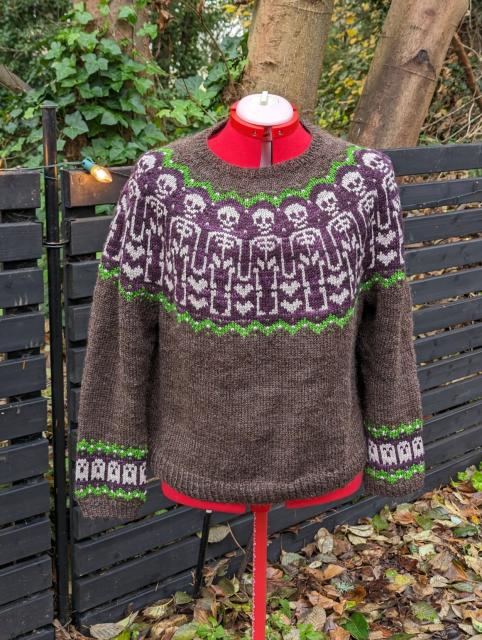 Finished object photo of a color work sweater, bone colored skeletons and ghosts on a purple and natural brown background.