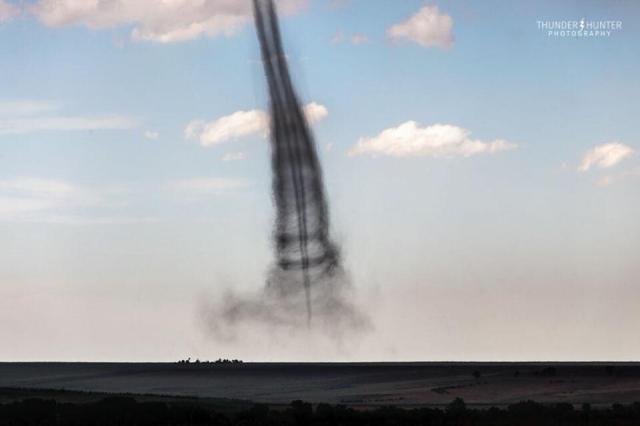 A funnel cloud is shown, but inside what appears to be a wider funnel cloud. A blue sky with a few white clouds is seen in the background, while flat plains are seen in the foreground.