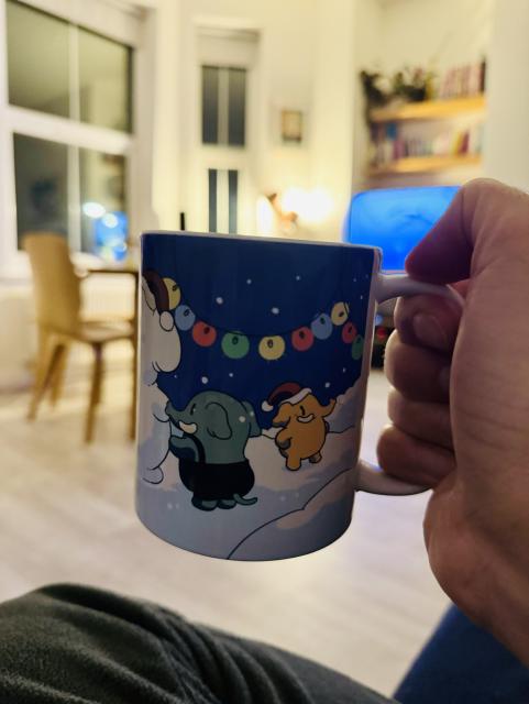 A hand holding a Mastodon mug with snow, balloons hanging, small cute mastodons playing. A living room is visible in the background.