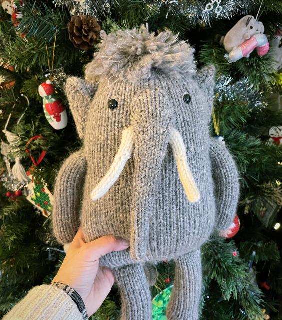 a photo of a knitted woolly mammoth doll, made from fuzzy taupe-coloured yarn. It is being held in front of a christmas tree with lights and ornaments