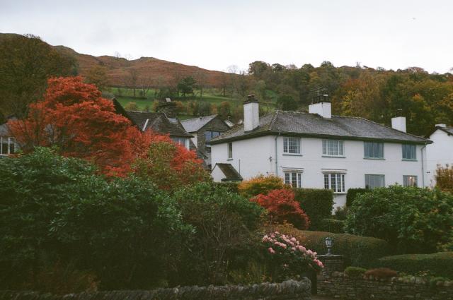 A white house with a grey roof and multiple chimneys, with a hill behind it. In front, there are green bushes and red trees, and in front of those, there are pink flowers.
