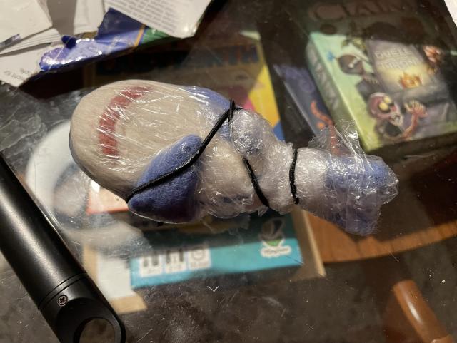 A felted shark covered in plastic wrap and secured with an elastic band, lying on a table. 