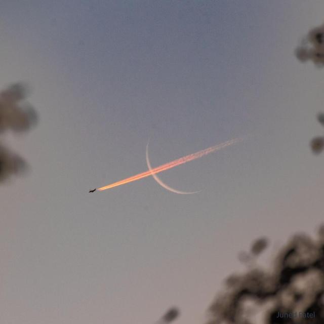 A thin crescent moon is shown with a bright red contrail going through it, right to left.