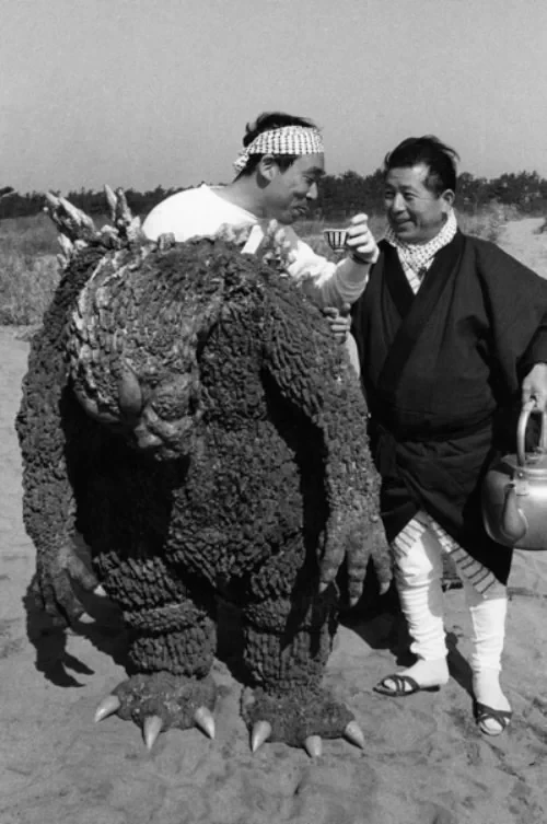 Picture of an actor in a partially undone Godzilla suit. The suit’s head and arms hang lazily down from the actor’s upper body as he drinks from a small cup of tea. A person in a dark colored poncho stands next to the actor holding an absolutely enormous tea pot. The thing is unhinged. It’s absolutely enormous. 