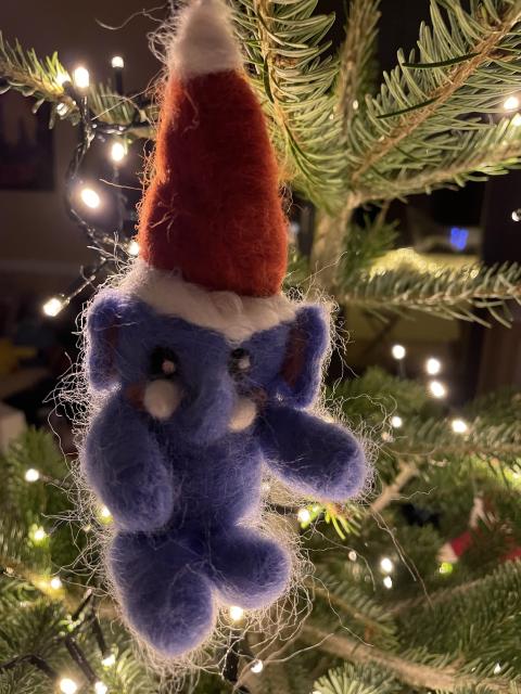 A blue felt Mastodon with rosy blushing cheeks in a red Santa hat hanging from a natural Christmas tree illuminated by pleasantly warm white fairy lights. 