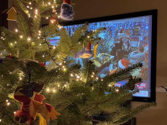 Natural Christmas tree with a blue felt Mastodon in a Santa hat bauble, a felt Blahaj in a Santa hat bauble, a plush Dota 2 Phoenix toy, a somewhat obscured felt donkey in a Santa hat bauble, and a ceramic Grasmere Gingerbread bauble facing the other way, illuminated by warm white fairy lights. In the background, a projector screen showing the Yogscast Jingle Jam livestream, currently on office cam. 