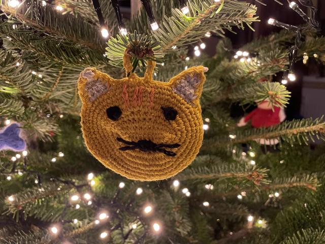 A yellow flat crochet cat hanging on a Christmas tree illuminated by warm white fairy lights. 