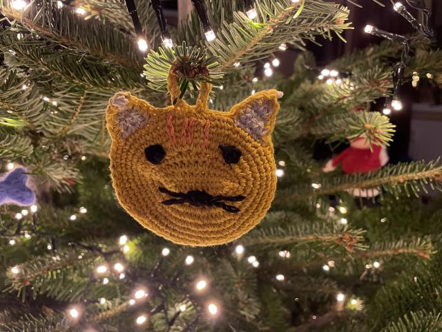 A yellow flat crochet cat hanging on a Christmas tree illuminated by warm white fairy lights. 