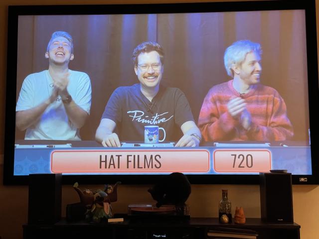 A projector screen showing the Jingle Jam livestream with the Hat Films game show program. On the frame, Ross, Alsmiffy and Trott can be seen. In front of Smiffy is the Mastodon winter mug. Below the projector screen you can also see a black cat, as a treat. 