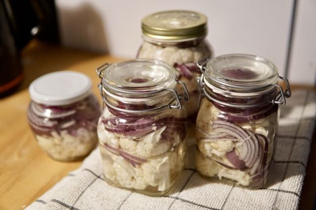 Various types of jars standing on a kitchen counter, all filled to the brim with cauliflower and red onion in brine.