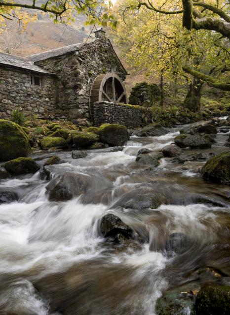 A stone built watermill stands beside a fast flowing stream. The stream is covered by autumnal trees. The water in the stream is slightly blurred by a long exposure as it cascades over rocks.