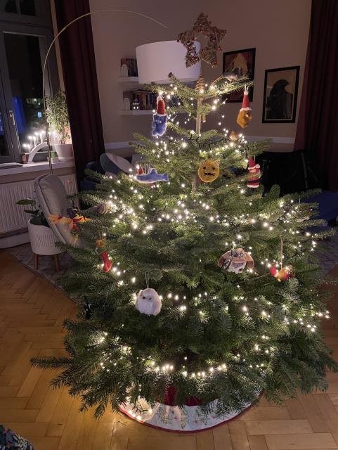 A living room with a green Christmas tree with warm white fairy lights, and a multitude of felt wool baubles as well as some crochet and plush ones, topped by a copper star. A red and white cover is underneath it. 