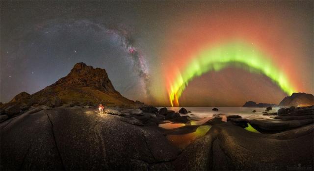 A night sky filled with stars is shown behind a picturesque foreground. The foreground contains rounded rocks and a person before a distant sea. The background contains bands of the Milky Way and bright aurora.