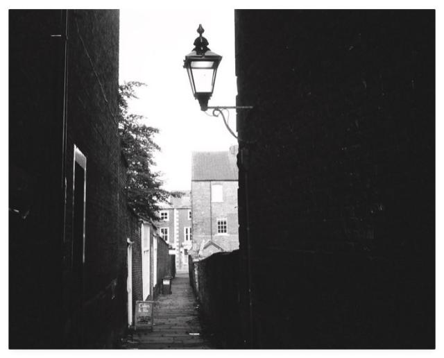 Gloomy black and white film photograph showing a shadowy alleyway with an old coaching lamp affixed to the right hand wall.