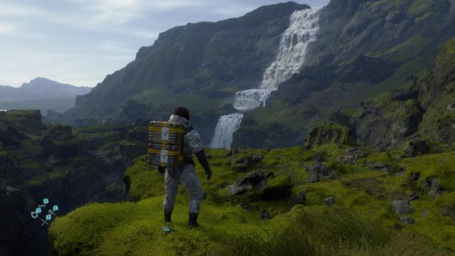 Screenshot of Sam from Death Stranding carrying items while looking at a waterfall