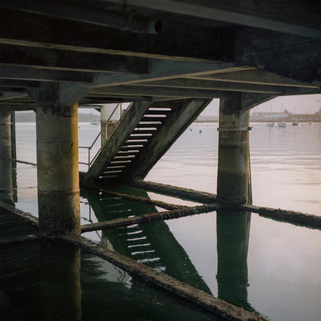 Underneath a concrete pier. Steps lead down to sea level. Reflections of the structure can be seen on the surface of the calm water.