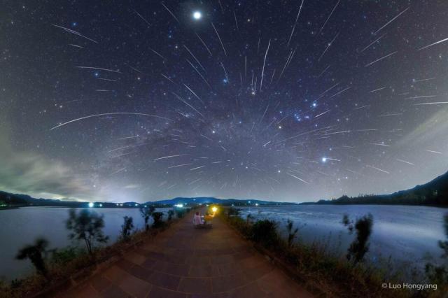 Two people are pictured from the back looking at a dark star-filled sky. The sky is also filled with numerous streaks caused by meteors from the Geminids meteor shower.