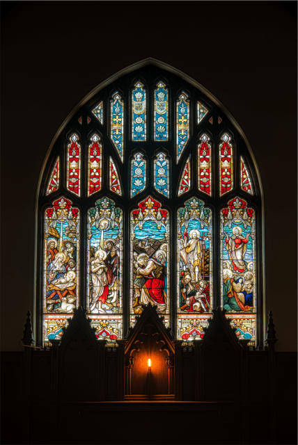 This image features a large, ornate stained glass window, commonly found in churches. The window is composed of several panels, each filled with vibrant colors and depicting what appear to be biblical scenes or figures. The intricate designs include reds, blues, greens, and yellows, with light shining through to highlight the details of each pane.

The window is Gothic in style, as suggested by the pointed arches at the top of each panel. Below the window, there's a dark interior space that contrasts with the bright glass, and a single lit candle or light source is positioned in the center at the bottom, casting a warm glow and creating an ambiance of reverence and spirituality.