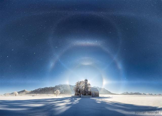 A building is seen from a distance on white snow and with mountains in the background. An ice-crystal filled sky is seen above. Superposed on the night sky are numerous curving whisps -- halos of ice reflecting background moonlight.